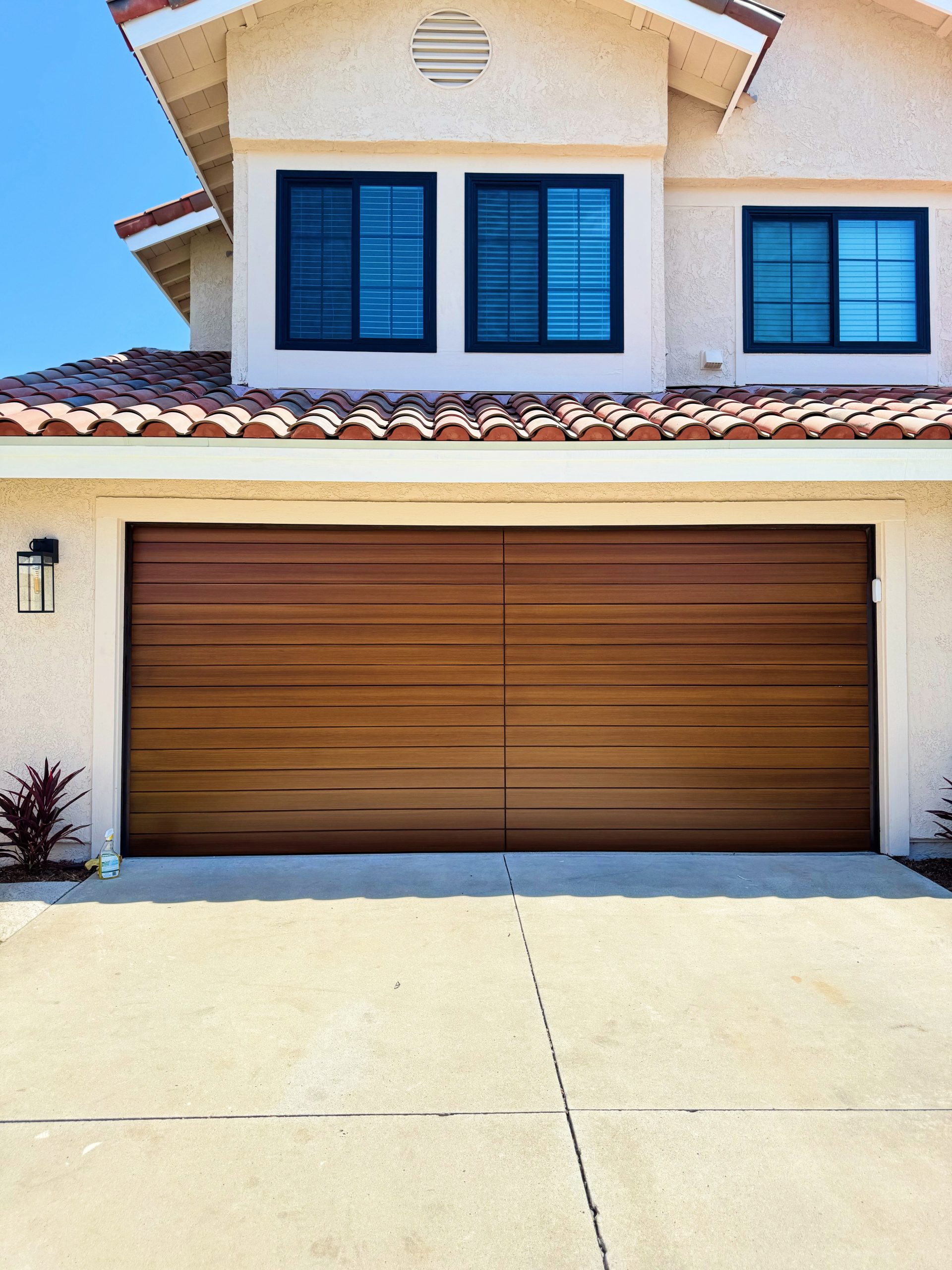 Modern garage door with a wood-like finish, featuring a sleek design, situated beneath a house with a tiled roof and black-framed windows, reflecting local architectural styles in Santa Ana, CA.