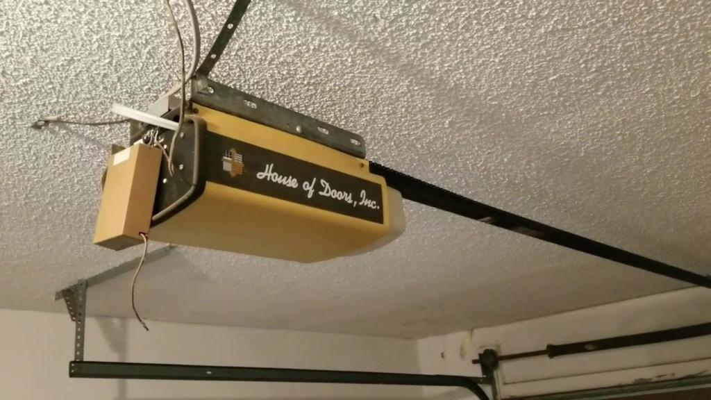 Garage door opener mounted on the ceiling, labeled "House of Doors, Inc.," illustrating an older model potentially in need of an upgrade for improved reliability and performance.